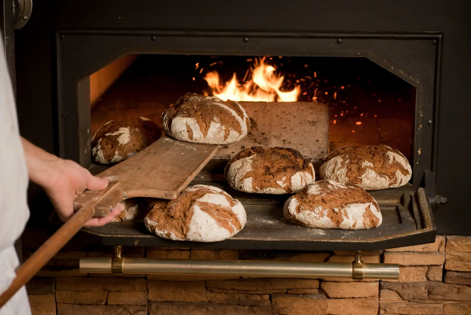 Ein Bäcker entnimmt rustikale Brotlaibe mit einem Holzschieber aus einem traditionellen Steinofen mit offenem Feuer
