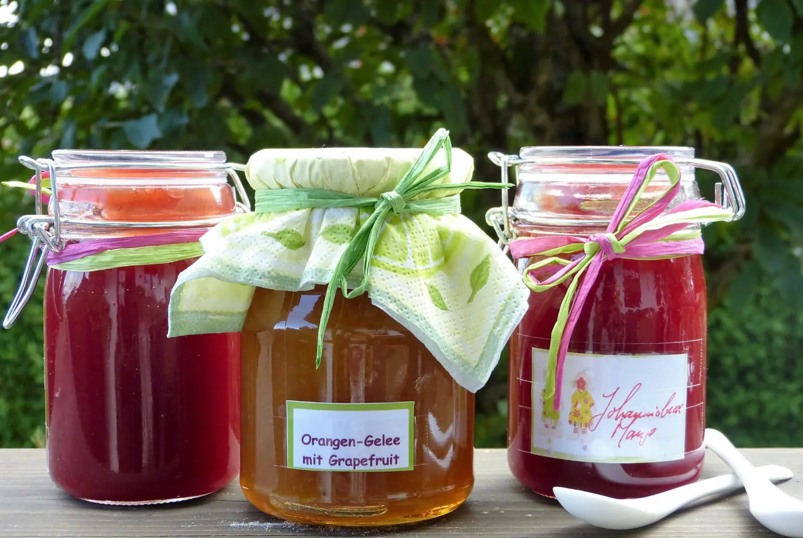 Drei dekorierte Einmachgläser mit hausgemachtem Gelee und Marmelade stehen auf einem Holztisch im Freien