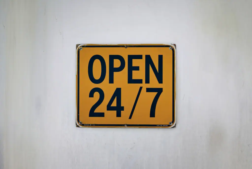 Gelbes quadratisches Schild mit der schwarzen Aufschrift „OPEN 24/7“ auf einer hellen Wand