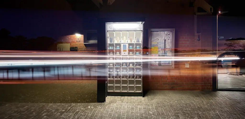 Beleuchtete marktbox bei Nacht mit Keramikprodukten und Lichtspuren vorbeifahrender Autos vor einer Backsteinwand