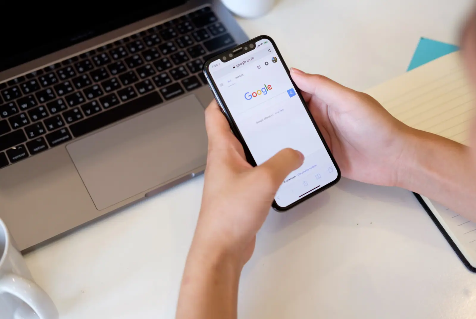 Hände bedienen ein Smartphone mit der Google-Suche neben einem Laptop und einem Notizbuch