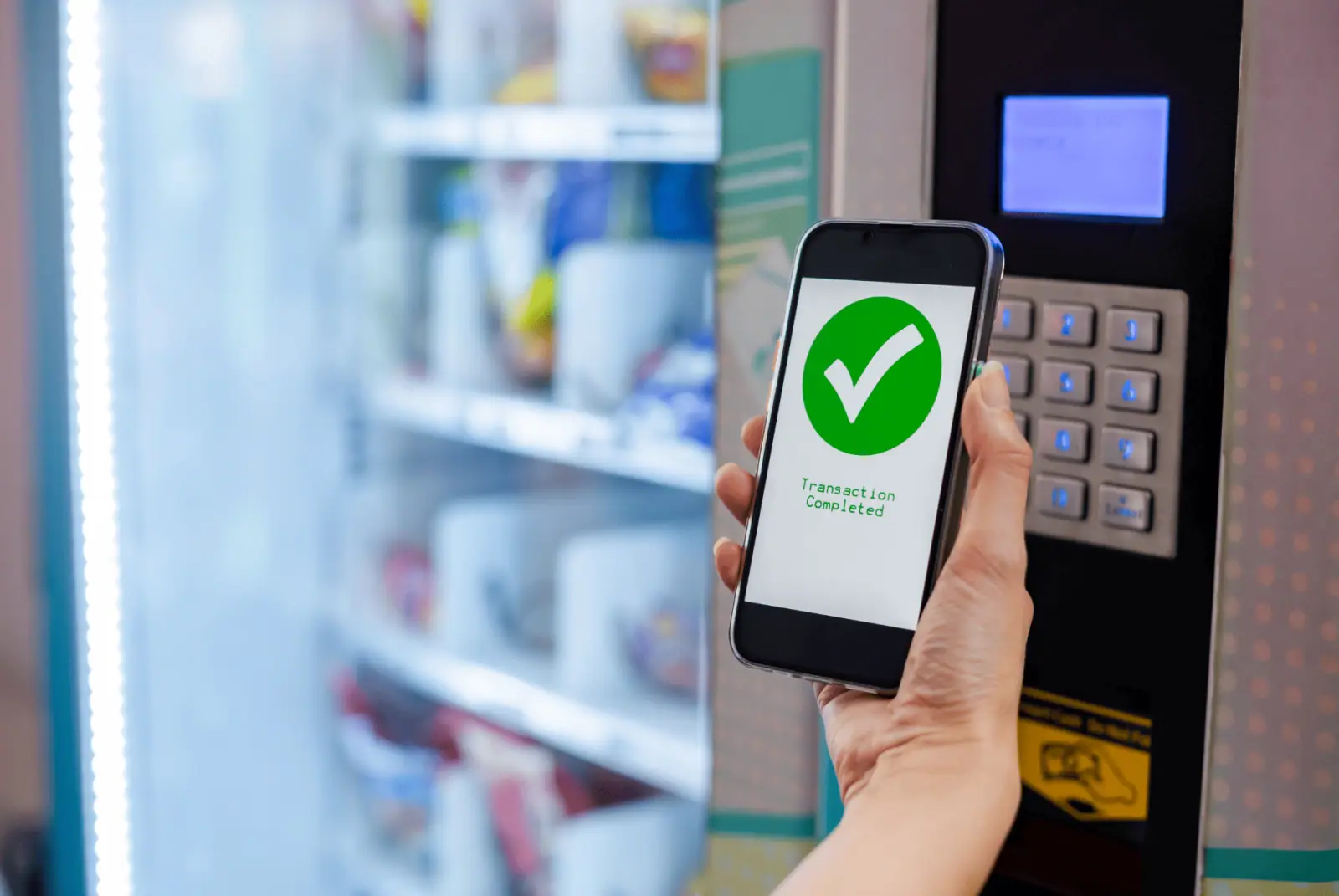 Hand hält ein Smartphone mit grünem Bestätigungshaken vor das Bedienfeld eines Verkaufsautomaten