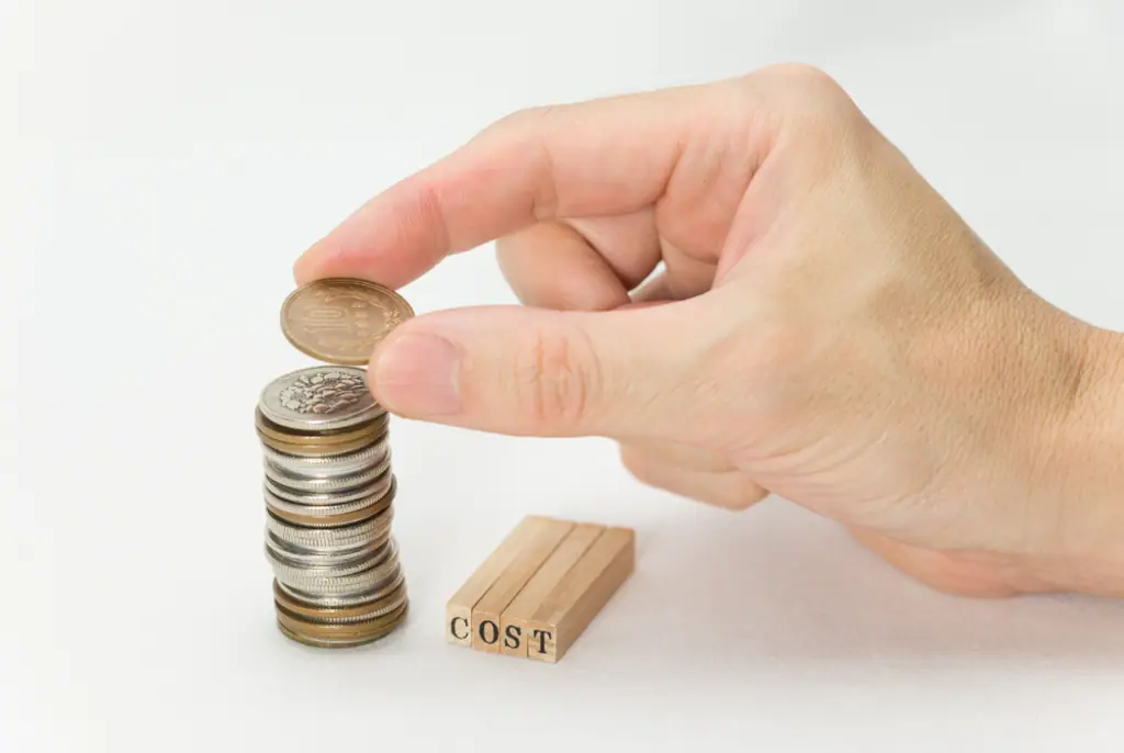Eine Hand legt eine Münze auf einen Stapel Geldmünzen neben Holzblöcken mit der Aufschrift COST