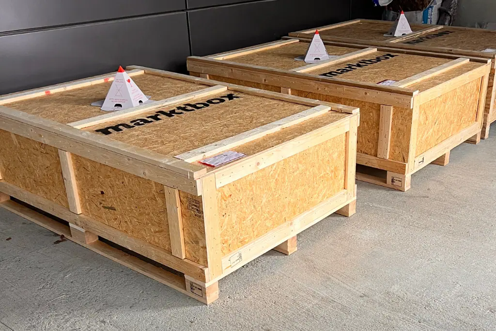 Drei große Holzkisten mit dem marktbox-Logo stehen versandfertig auf einem hellen Betonboden