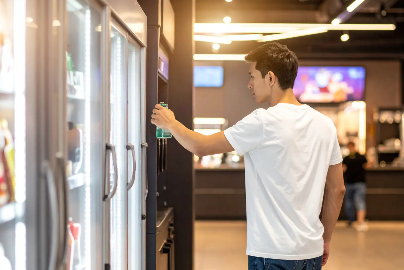 Junger Mann entnimmt eine Glasflasche aus einem modernen Getränkeautomaten mit beleuchteter Glasfront