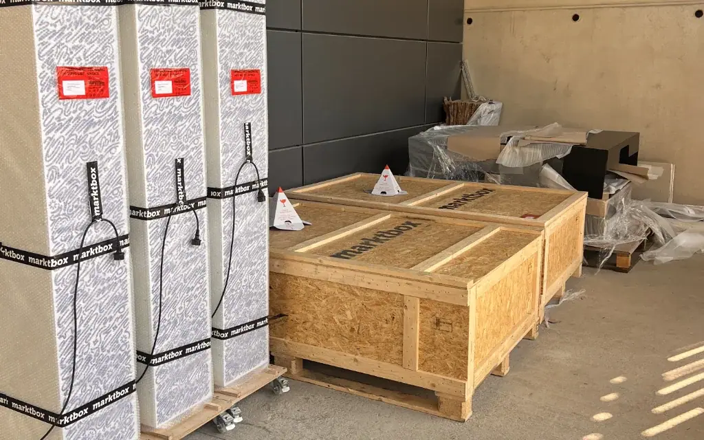 Drei hochkant verpackte Marktboxen und zwei große Transportkisten aus Holz in einer Produktionshalle