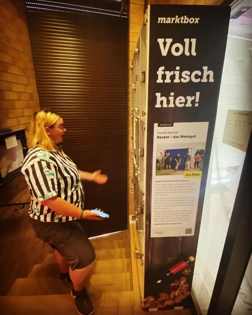 Nutzerin bedient einen marktbox-Verkaufsautomaten mit der Aufschrift Voll frisch hier über ihr Smartphone