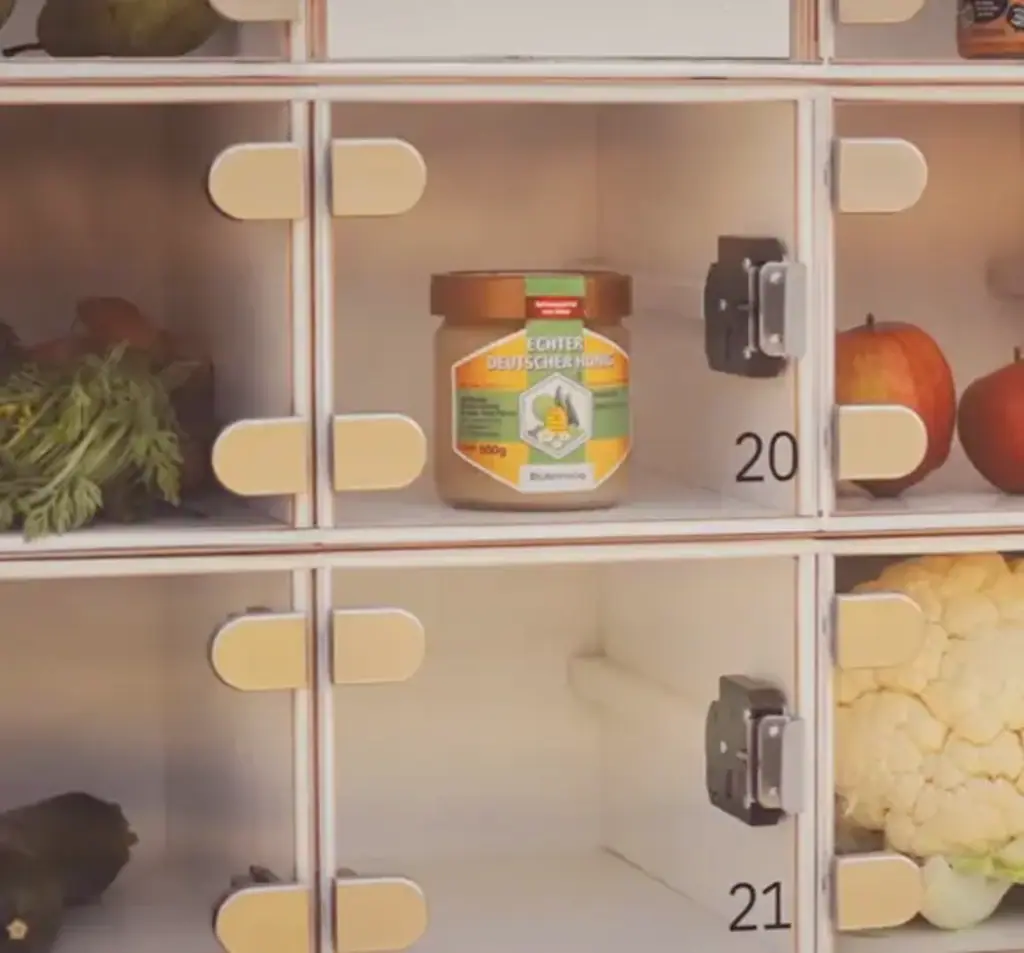 Fächer eines Klappenautomaten gefüllt mit einem Glas Honig, Äpfeln und frischem Gemüse