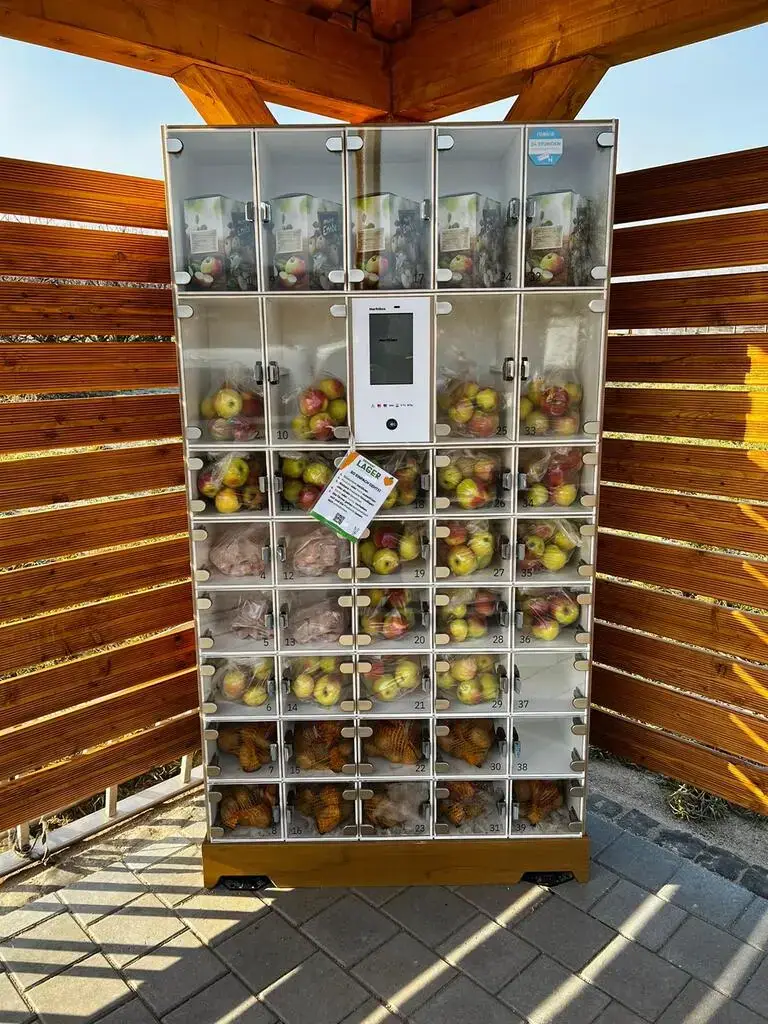 Marktbox-Verkaufsautomat mit Glasfächern für Äpfel, Kartoffeln und Saft in einem hölzernen Unterstand