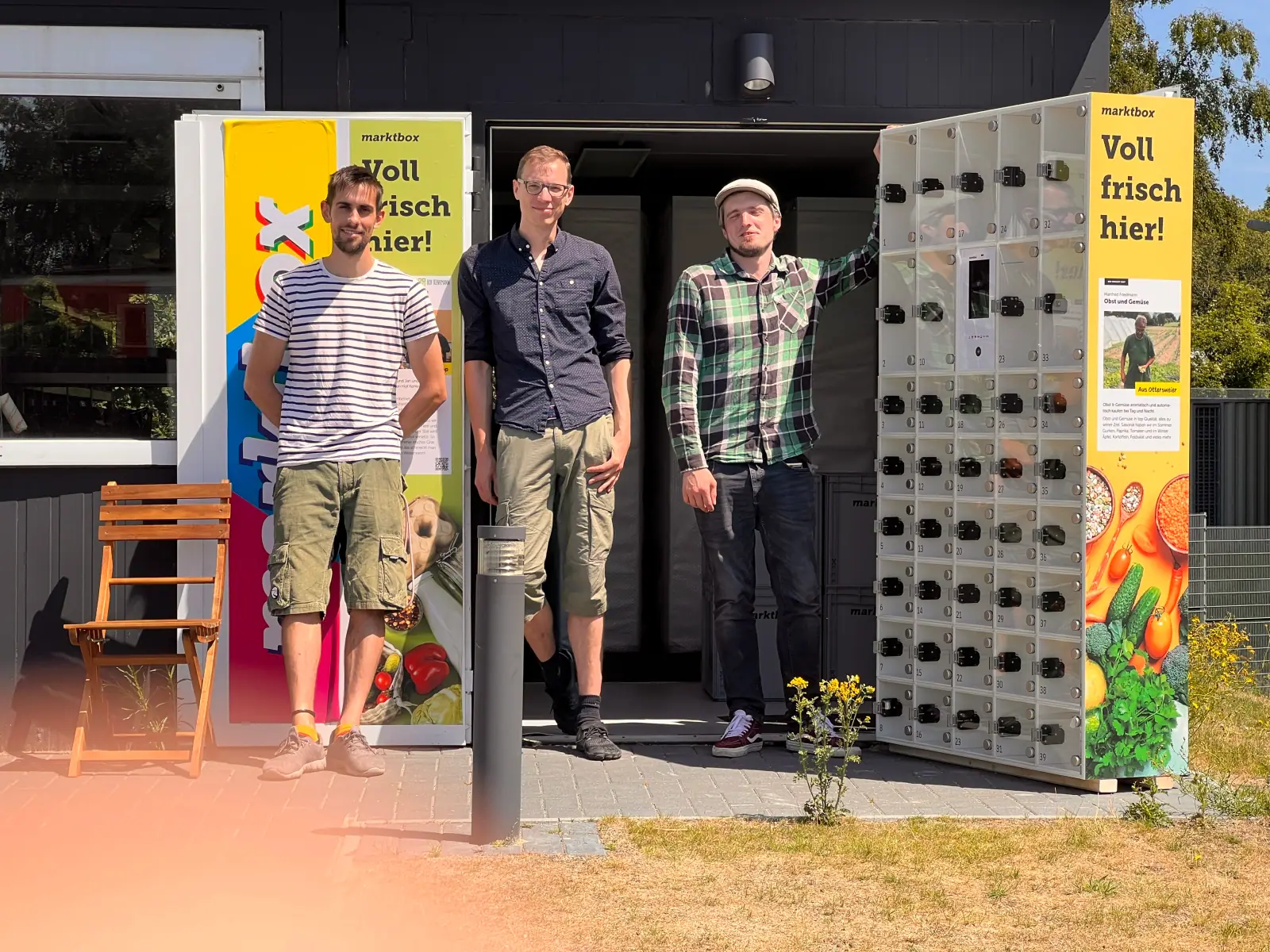 Drei Männer stehen vor einer gelben Marktbox mit Schließfächern und digitalem Terminal im Freien