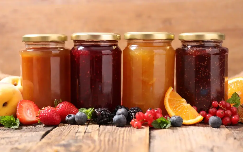 Vier Gläser mit verschiedenen Konfitüren auf einem Holztisch neben frischen Erdbeeren, Beeren und Orangen